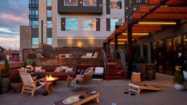 Outdoor patio area with seating, fire pit, string lights, cornhole boards, and modern building in the background at sunset.