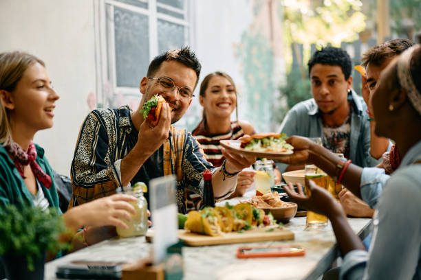 A group of young adults sit around an outdoor table, eating tacos and sandwiches, smiling and enjoying drinks together in a casual setting.
